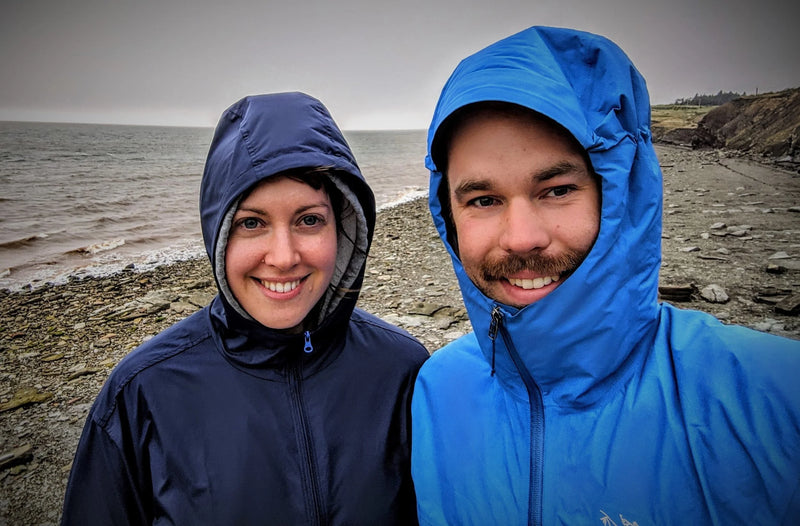 Two people wearing raincoats standing on a rocky shoreline with water in the background.