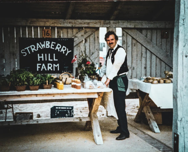 Man standing behind a table with produce in a barn setting, featuring 'Strawberry Hill Farm' sign.