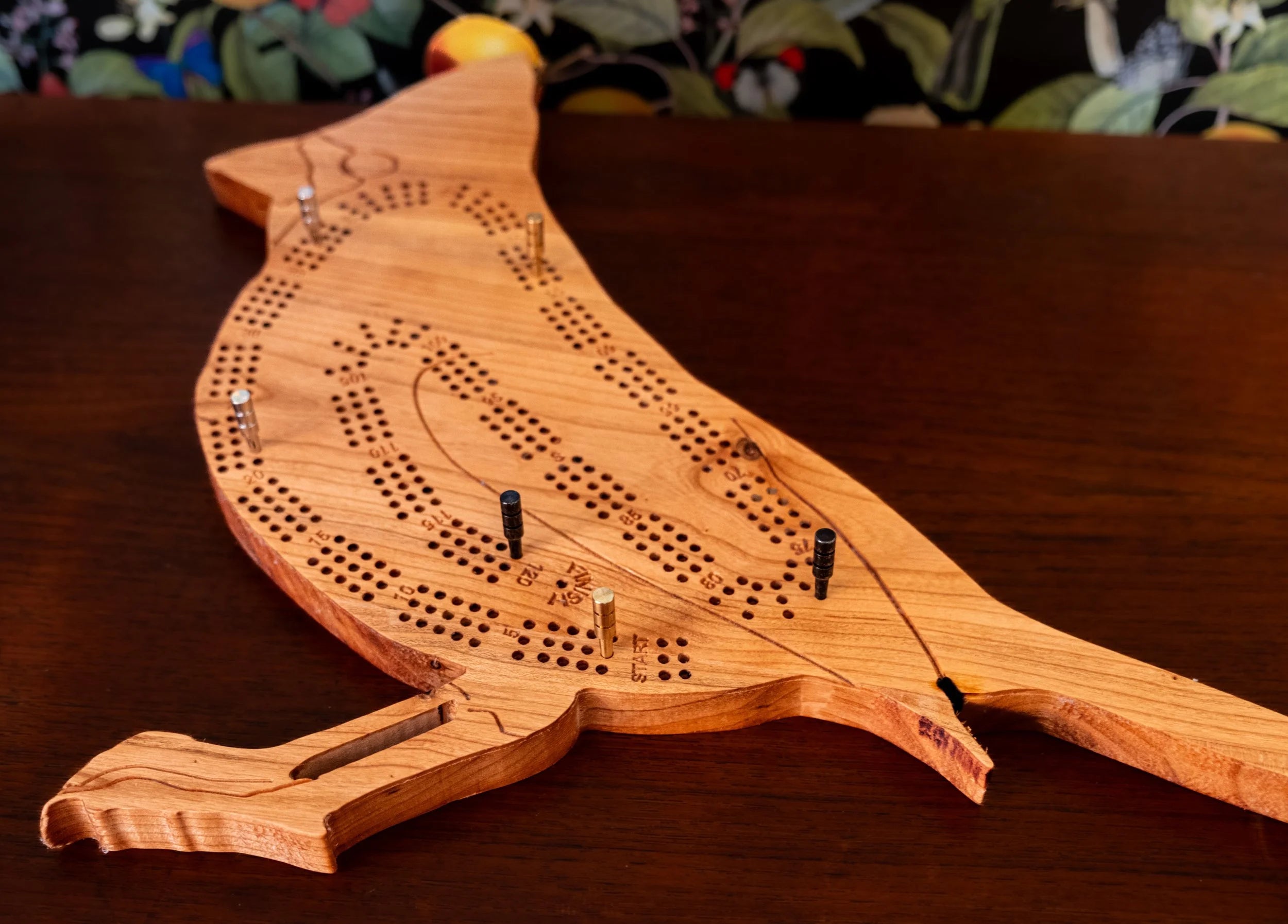 Wooden bird-shaped cribbage board on a wooden surface with a floral background.