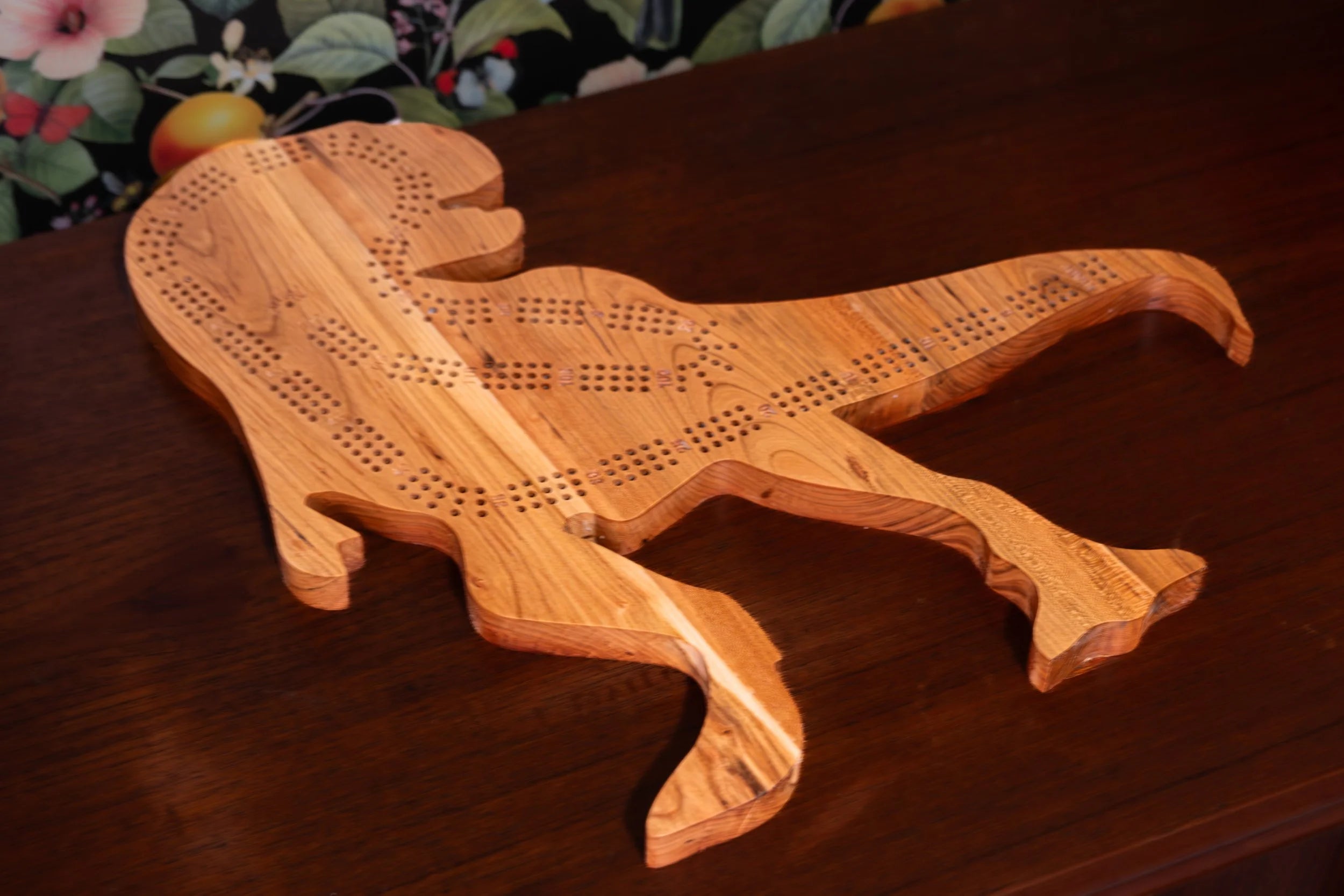 Wooden t-rex dinosaur shaped cribbage board on a wooden surface with a floral-patterned fabric background.