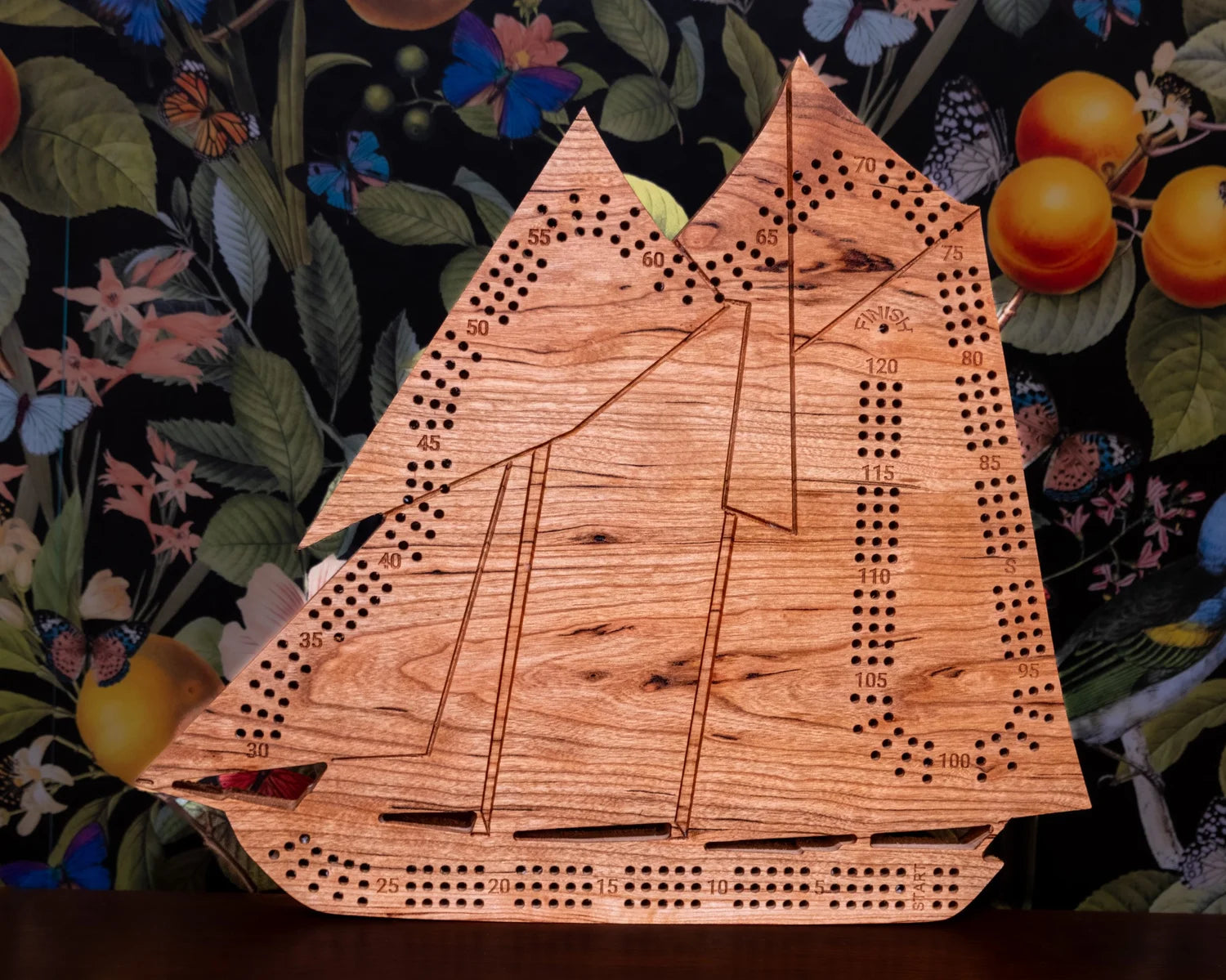 Wooden ship-shaped cribbage board on a floral background with fruits.
